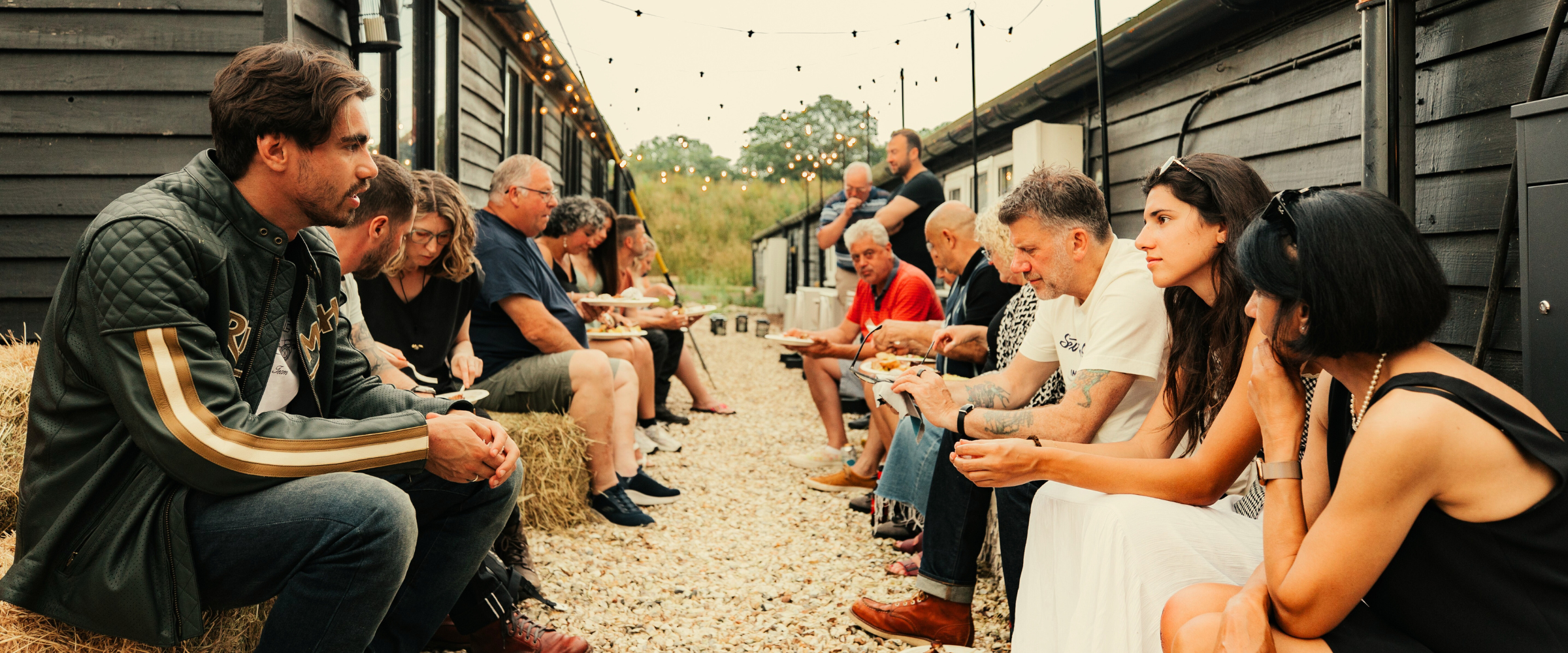 Motorcyclists and guests sharing food and stories at a Machinehead Moto social evening, seated outdoors between workshop cabins with festoon lights above.
