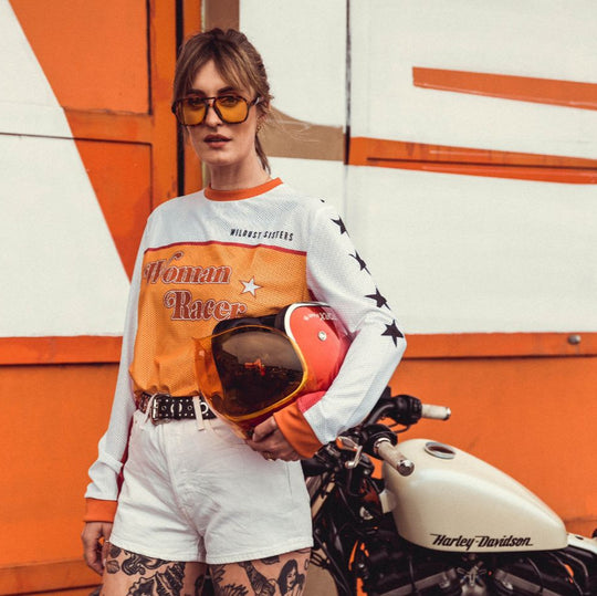 Model holding motorcycle helmet, wearing Wildust Sisters Jersey Cross – Woman Racer, with Harley Davidson motorcycle
