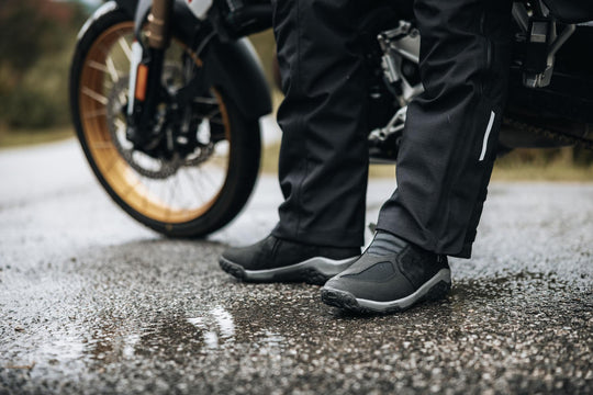 Boundless H2O ADV Footwear worn beside an adventure motorcycle on a wet road.
