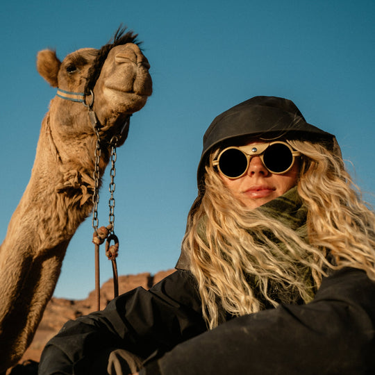 Person with long blonde hair and sunglasses standing next to a camel in a desert setting