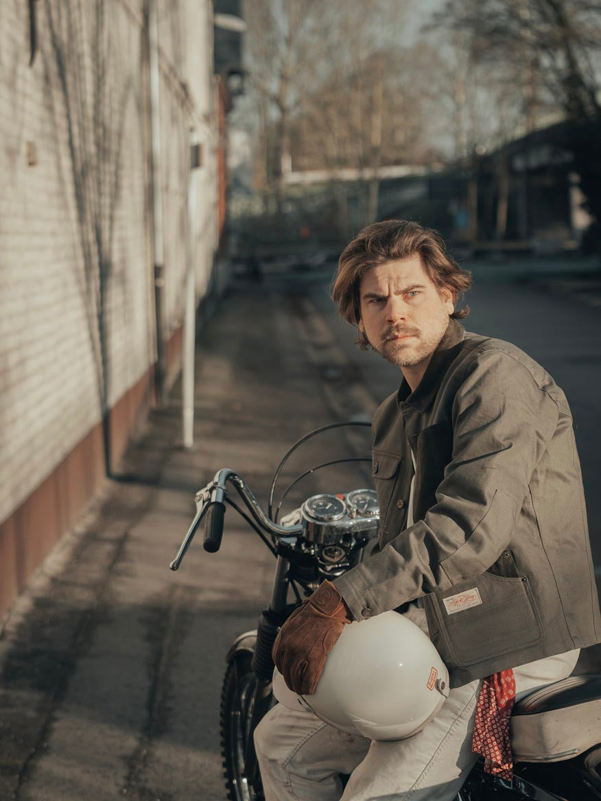 Model Wearing Age Of Glory Craftsman Jacket in Olive, 3/4 View, Buttoned cuffs and Pockets sat on Motorcycle
