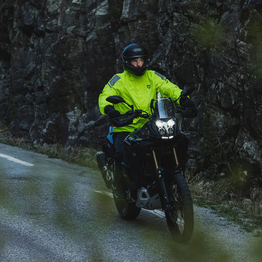 Model riding a motorcycle in wet conditions wearing a high-vis REV’IT! rain jacket on a winding road