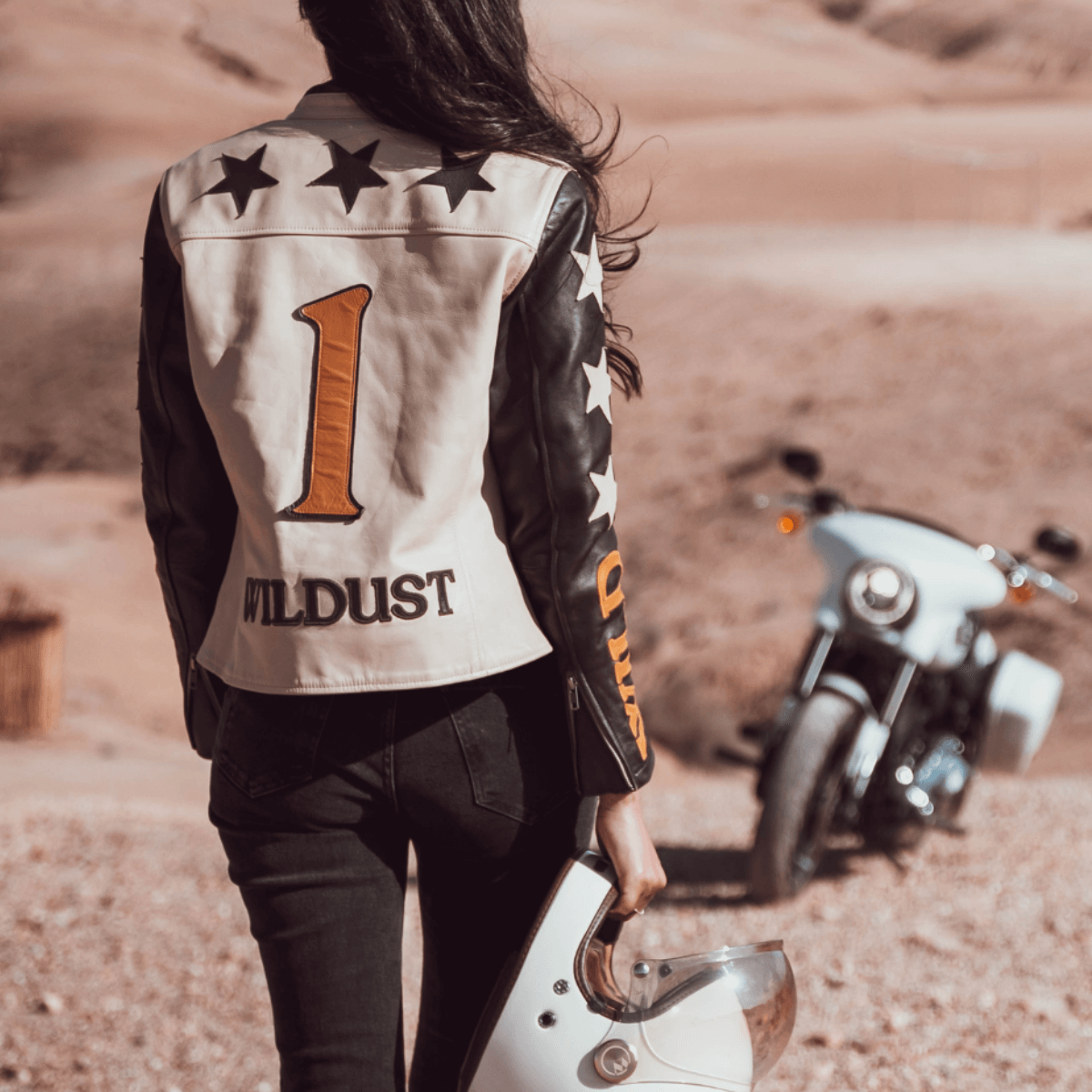 Woman in a Wildust 'Stars and Stripes' leather jacket holding a helmet, standing in a desert landscape with a white motorcycle in the background.
