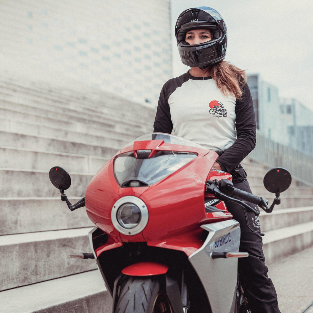Rider wearing the Wildust Sisters Rebel Long Sleeve Tee with black sleeves and red helmet graphic, seated on a red motorcycle outdoors.
