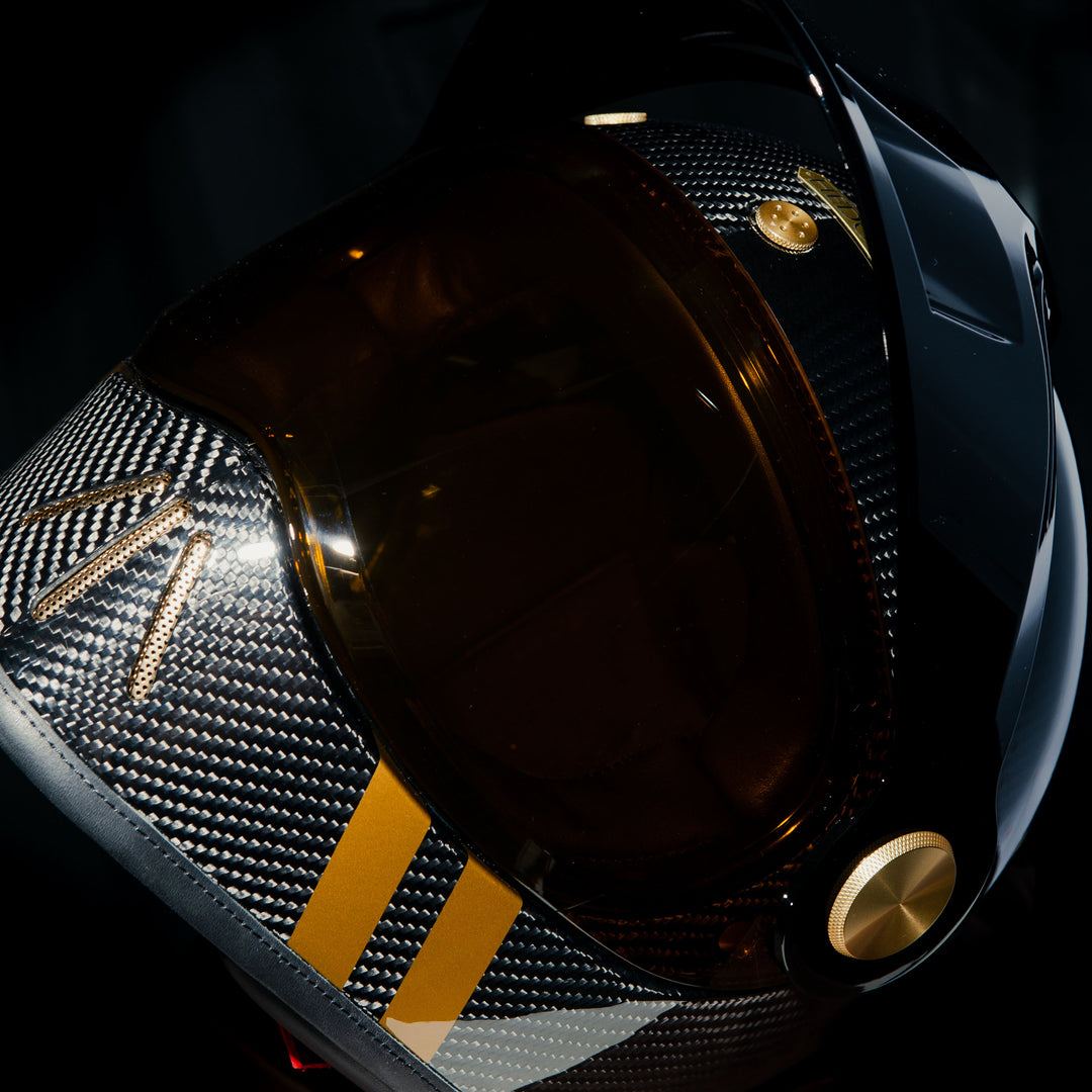 Close-up angled view of the Hedon Psilo Explorer Helmet – Commander with the tinted visor closed, showcasing the exposed carbon fibre shell, gold visor hardware, and signature gold chin stripes.