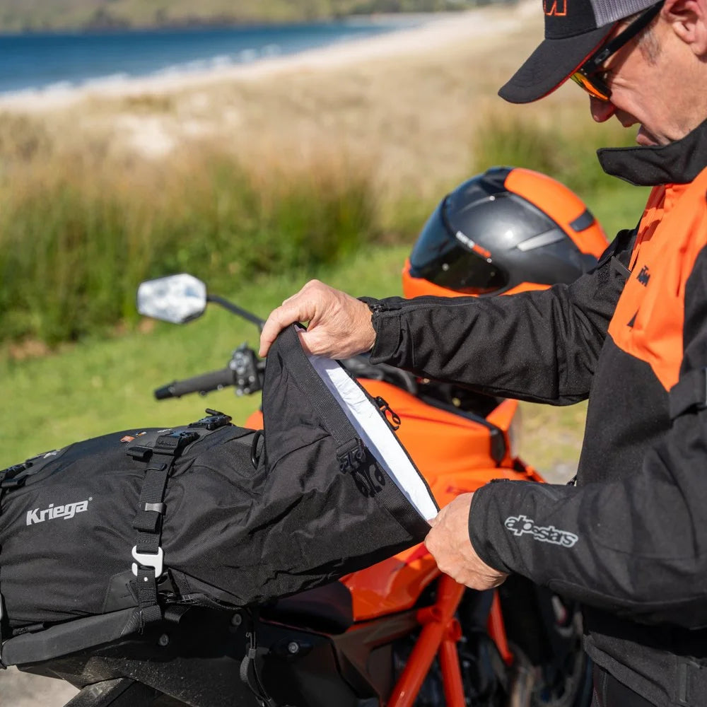 Rider adjusting Kriega US-20 Drypack mounted on an orange KTM motorcycle near a lake