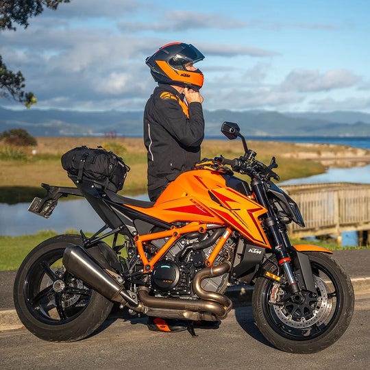 Kriega US-20 Drypack mounted on an orange KTM motorcycle, rider in helmet with scenic lake and hills in the background