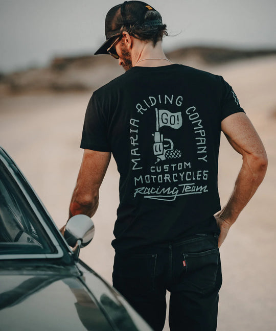Man wearing a black t-shirt with motorcycle branding, standing next to a car in a desert setting.