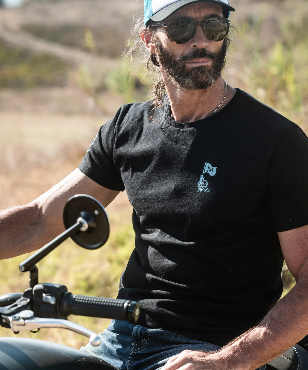 Man wearing a black t-shirt with a logo, sitting on a motorcycle in an outdoor setting.