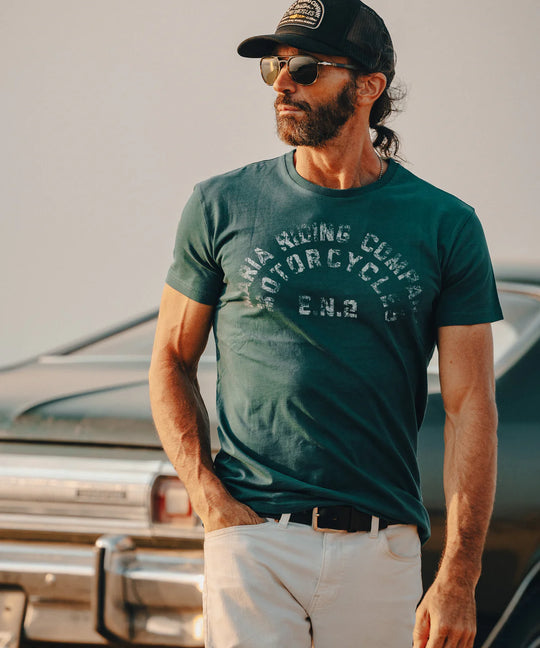 Man wearing a green t-shirt with text, sunglasses, and a cap standing in front of a vintage truck.