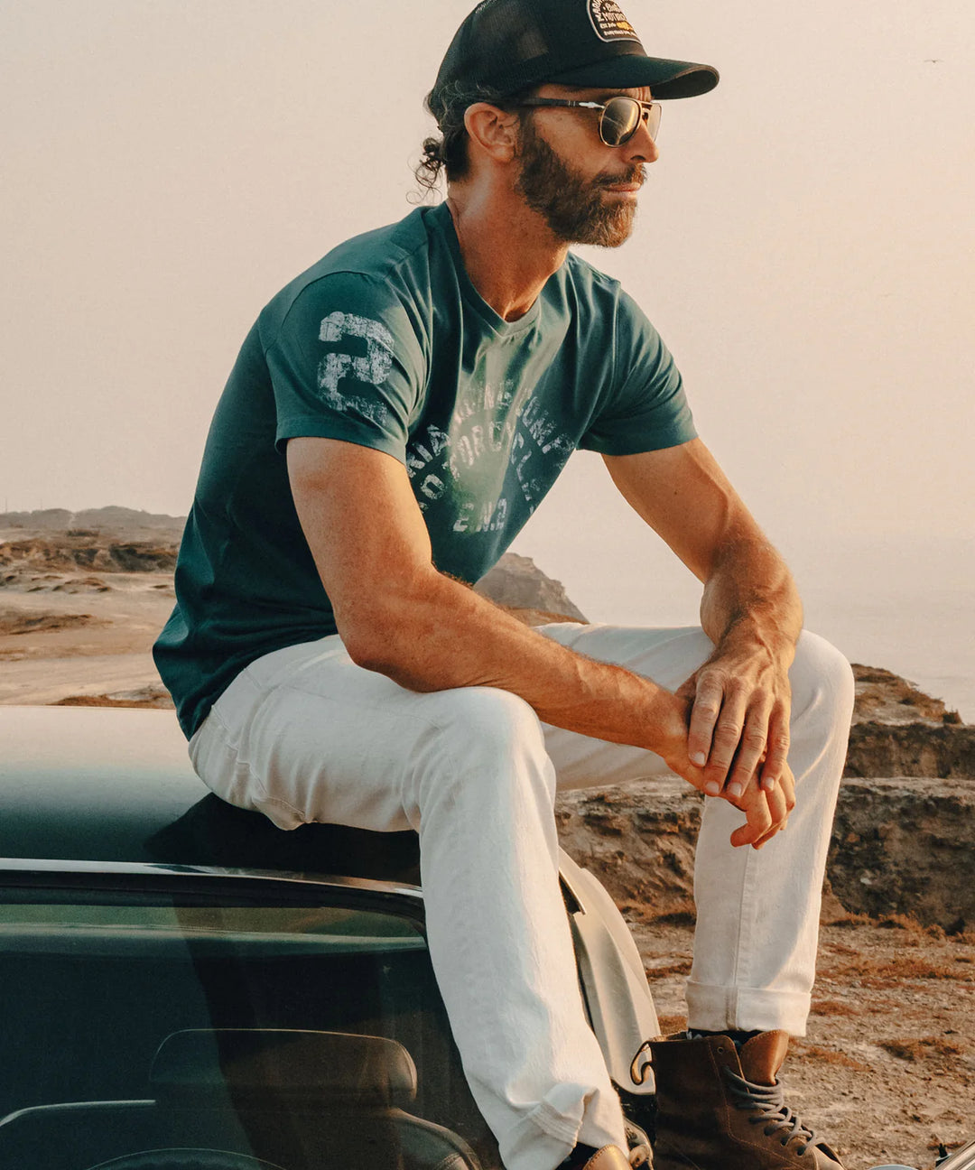 Man sitting on a car wearing a green t-shirt, white pants, and a black cap with sunglasses on a scenic background.