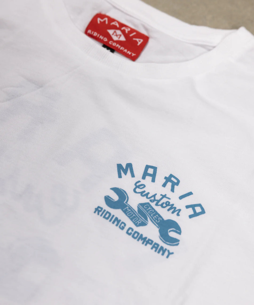 White t-shirt with blue 'Maria Custom Riding Company' logo and red tag on a beige background