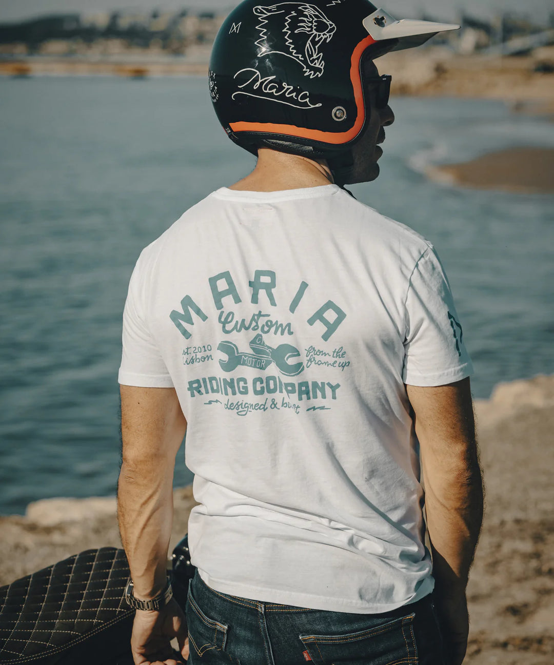 Person wearing a white t-shirt with 'Maria Custom Riding Company' text, sitting by a body of water.