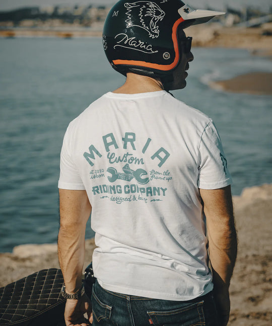 Person wearing a white t-shirt with 'Maria Custom Riding Company' text, sitting by a body of water.