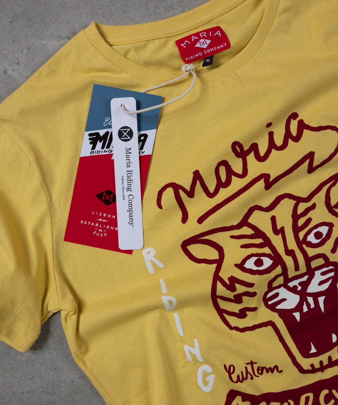 Yellow t-shirt with red tiger graphic and 'Maria Riding' branding on a gray surface.