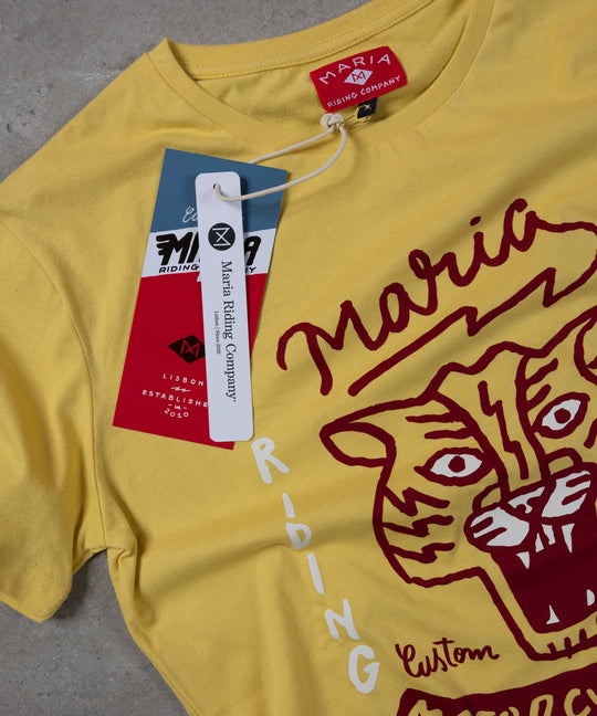 Yellow t-shirt with red tiger graphic and 'Maria Riding' branding on a gray surface.