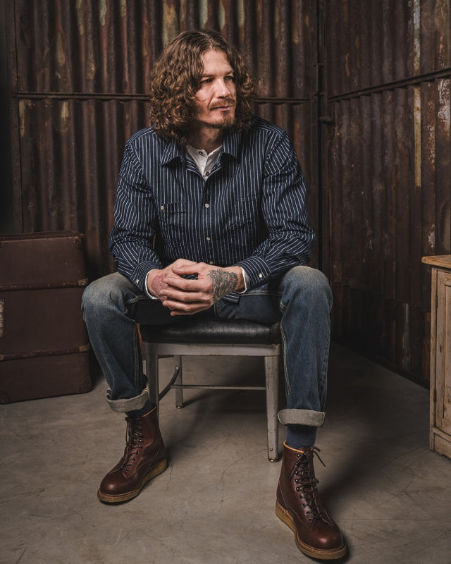 Man seated wearing the Pike Brothers 1937 Roamer Shirt in blue Wabash denim, shown full outfit in a workshop-style setting.