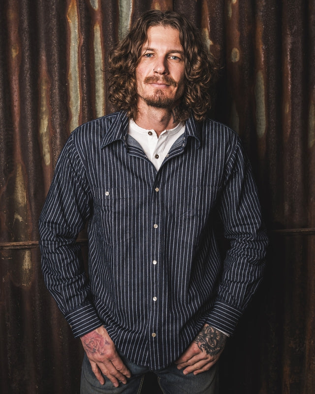 Man standing wearing the Pike Brothers 1937 Roamer Shirt in blue Wabash denim, front view against a metal backdrop.
