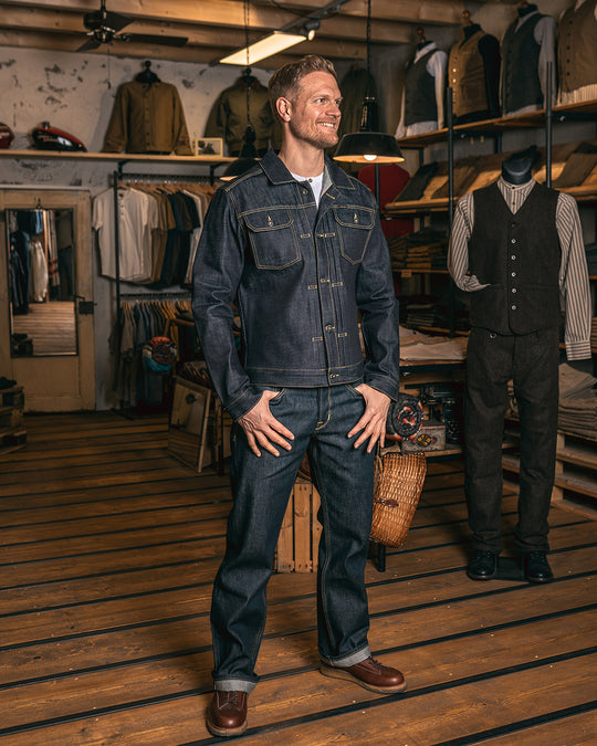 3/4 view of model wearing Pike Brothers 1958 Roamer Jacket in 15oz indigo denim with denim Chopper Pants, photographed in shop setting