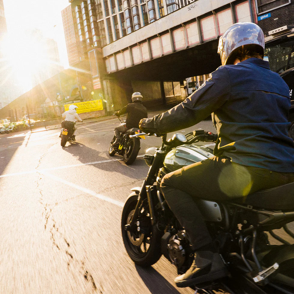 Model riding a motorcycle wearing REV’IT! jacket, trousers and Arrow 2 shoes, following two other riders through an urban street at sunset