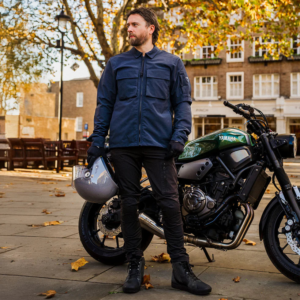 Model wearing a REV’IT! motorcycle jacket, black riding trousers and REV’IT! Arrow 2 shoes, standing beside a Yamaha motorcycle holding a helmet in an urban setting
