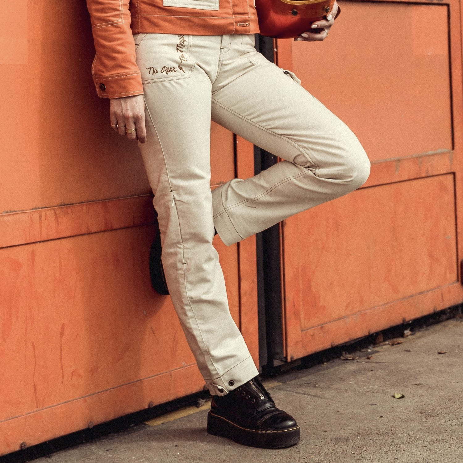 Female rider wearing Wildust Sisters Ivory Cargo Trousers with helmet, leg bent against orange container showing pocket embroidery