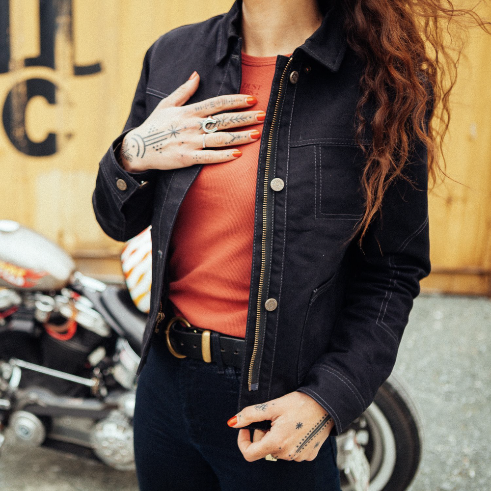 Close-up of female rider’s tattooed hand wearing Wildust Sisters Pioneer Jacket in Black, open at front with brick-coloured top and black jeans