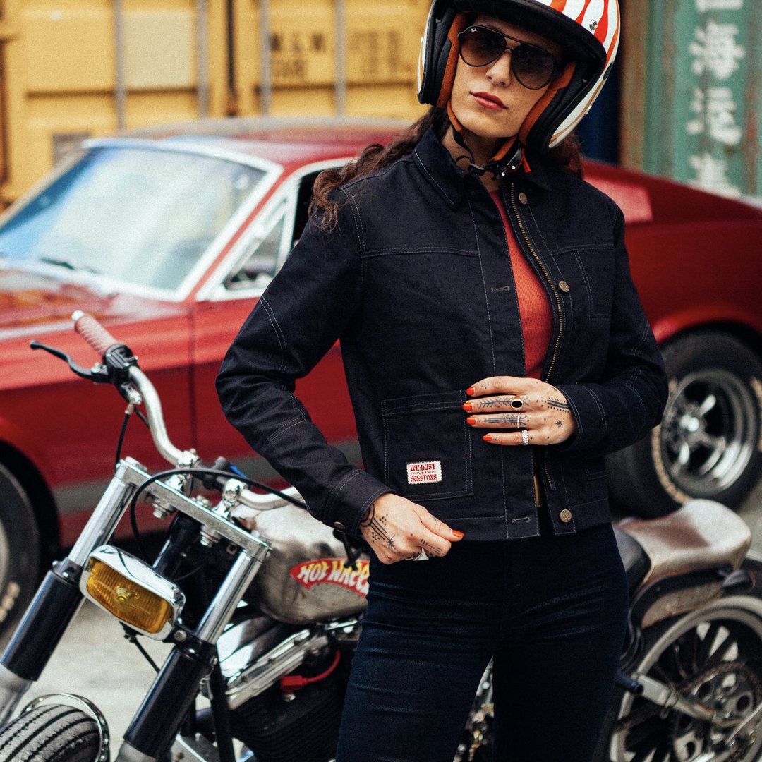 Female rider wearing Wildust Sisters Pioneer Jacket in Black with helmet and sunglasses, front view showing open zip pockets, contrast stitching, buttons, and pocket badge
