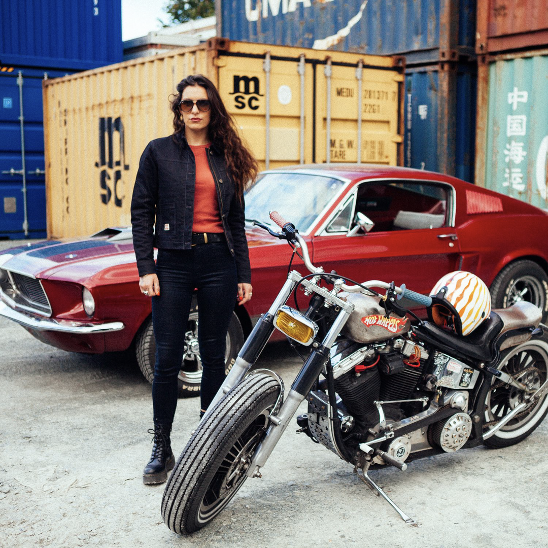 Female rider with long hair wearing Wildust Sisters Pioneer Jacket in Black, standing next to motorcycle and Dodge car with shipping containers, jacket open and sunglasses on