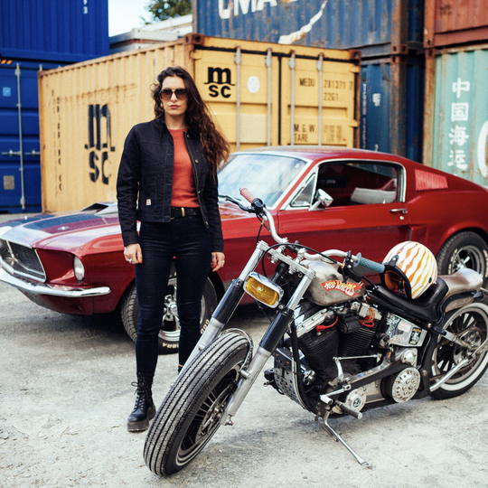 Female rider with long hair wearing Wildust Sisters Pioneer Jacket in Black, standing next to motorcycle and Dodge car with shipping containers, jacket open and sunglasses on