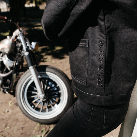 Close-up side view of female rider wearing Wildust Sisters Pioneer Jacket in Black showing “No Risk No Magic” embroidery
