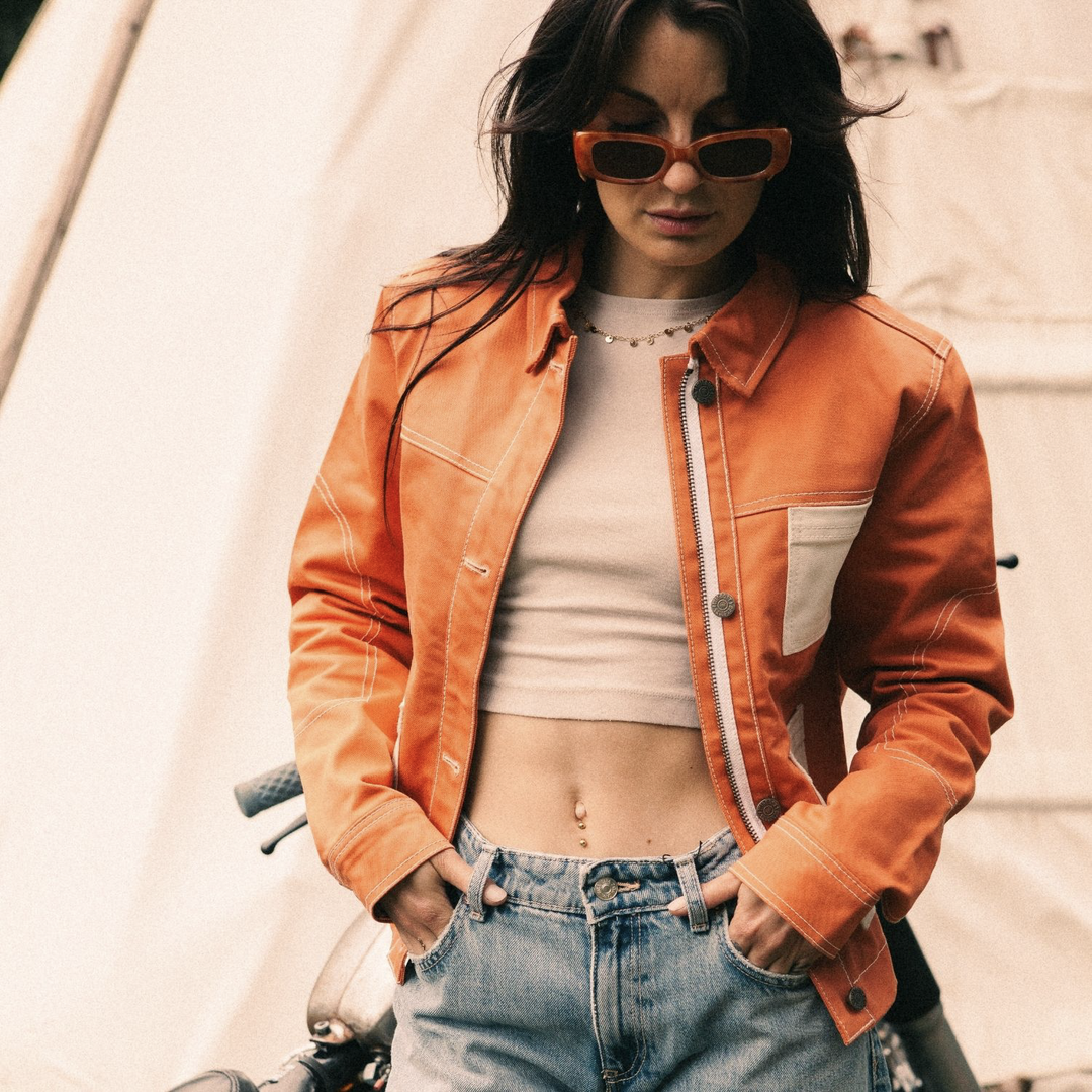 Female rider wearing Wildust Sisters Pioneer Jacket in Orange, unzipped with jeans and sunglasses, front view