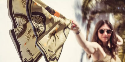 Rider holding up the Wildust Sisters Ride & Shine Scarf in khaki, ecru and orange, showing the full tattoo-inspired motorcycle print outdoors