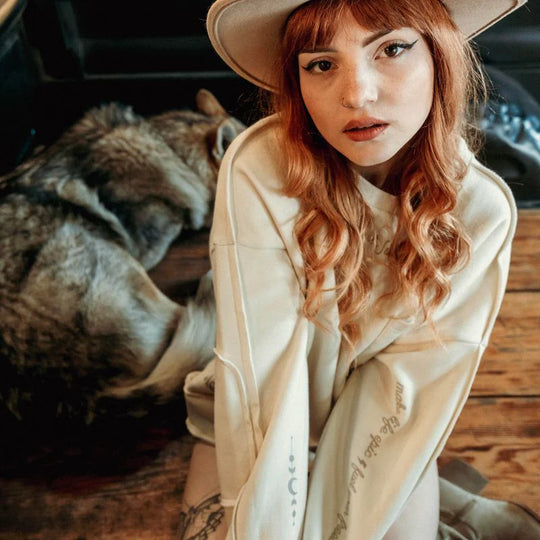 Model wearing the Wildust Sisters Escape Sweatshirt with sleeve prints and a beige hat, sitting indoors beside a wolf-like dog on wooden flooring.
