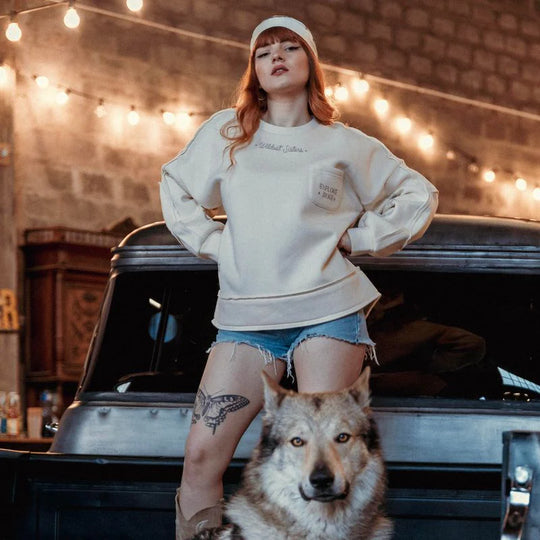Model wearing the Wildust Sisters Escape Sweatshirt with “Explore More” pocket print, standing beside a wolf in a workshop-style setting with warm lights.