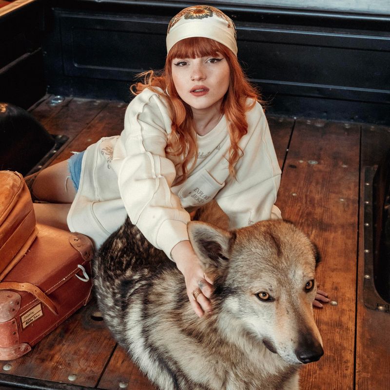 Model wearing the Wildust Sisters Escape Sweatshirt in ecru with “Explore More” pocket, sitting beside a wolf-like dog on wooden flooring with leather bag nearby.