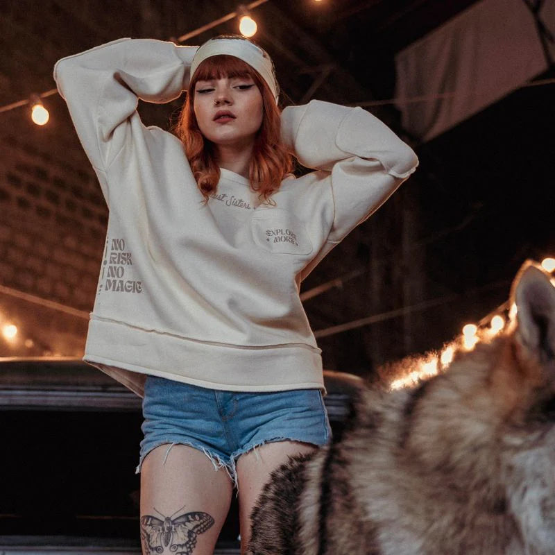Model wearing the Wildust Sisters Escape Sweatshirt in ecru with “Explore More” pocket and sleeve text, standing beside a wolf-like dog in a rustic indoor setting.