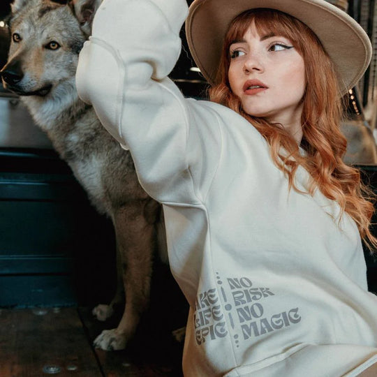 Model wearing the Wildust Sisters Escape Sweatshirt with “No Risk No Magic” side print, beige hat, and visible wolf in the background.