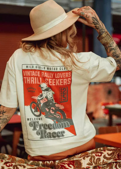 Model in hat wearing Wildust Sisters Freedom Racer T-shirt, showing large back graphic while seated.