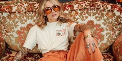 Model wearing Wildust Sisters Freedom Racer T-shirt with front chest logo, sunglasses, and orange trousers, seated on patterned sofa.